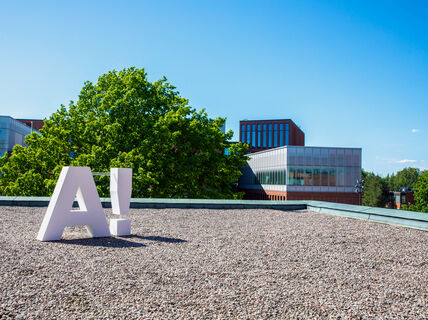 A large white 'A!' sculpture on the rooftop of the Undergraduate centre. A large tree and other buildings in the background.
