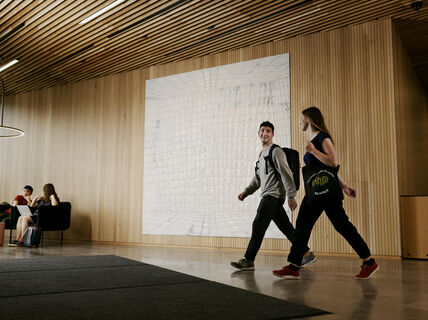 Two people walk through a modern lobby with a light wood interior and a geometric wall art. Others sit on black chairs.