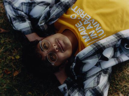 Person lying on grass wearing a yellow shirt with white text and a black-and-white checkered jacket.