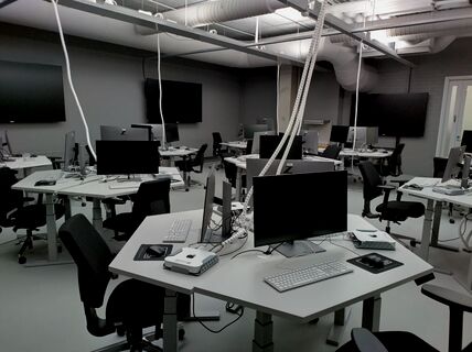 A modern computer lab with multiple workstations. Each desk has monitors, keyboards, and various devices connected.