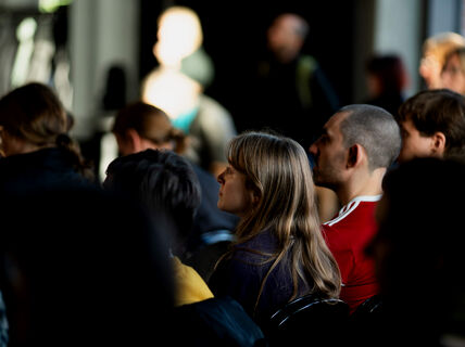 A group of people sitting indoors attending an event. The focus is on a person with long hair, wearing a dark top.