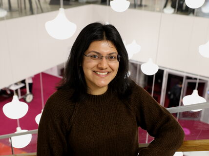 Person with black hair wearing a brown sweater, standing in a modern room with hanging lamps and glass railings.