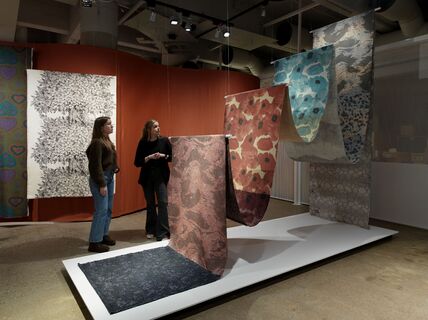 Two women observe a display of patterned fabrics hanging vertically in a showroom with an orange backdrop.