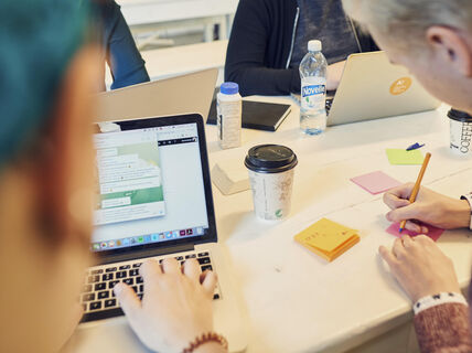 Ihmisiä työskentelemässä pöydän ääressä, kannettavat tietokoneet, muistilaput ja kahvikupit. Yksi henkilö kirjoittaa muistiinpanoja.