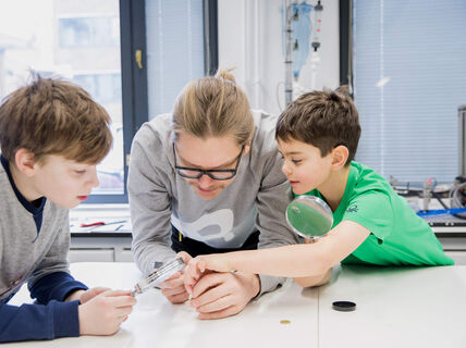 Three people are examining an object on the table in a well-lit room with large windows and scientific equipment.