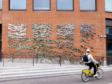 A person rides a yellow bike past a brick wall with abstract metal art installations. The rider wears a hat and has a backpack.