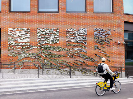 A person rides a yellow bike past a brick wall with abstract metal art installations. The rider wears a hat and has a backpack.