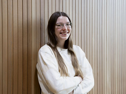 Person with long brown hair, wearing a white jumper, standing against a wooden panel wall, arms crossed.