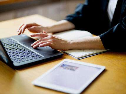 A person typing on a laptop next to a notebook and tablet on a wooden desk.