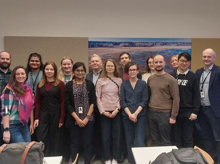 Group of eleven people standing in an office setting, some wearing ID badges. There's artwork on the wall behind them.
