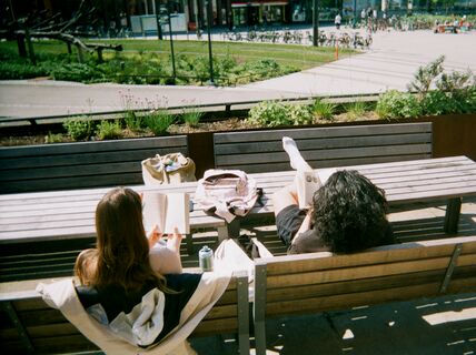 Two people are sitting on wooden benches outside, reading books. There is a grassy area and a bike park in the background.