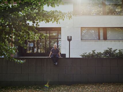 A man wearing a dark shirt sits on a concrete wall in front of a building with large windows and trees nearby.