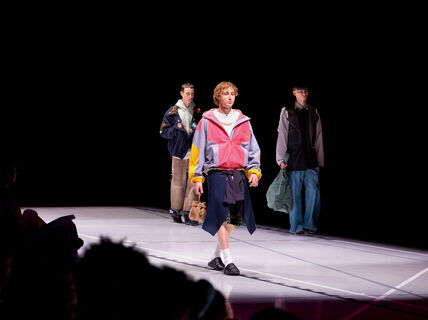 Three models walk on a runway. The front model wears a colourful jacket, black shorts, and black shoes.