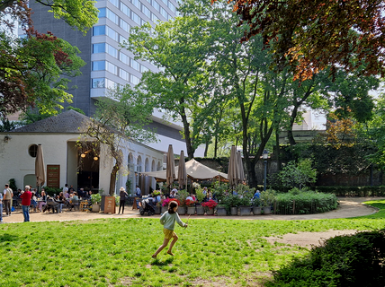 A girl runs on grass in a park with a café, trees, and people in the background. A tall building is visible behind the café.
