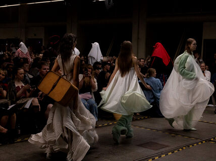 Fashion show with models in avant-garde outfits walking down the runway, audience seated on both sides taking photos.