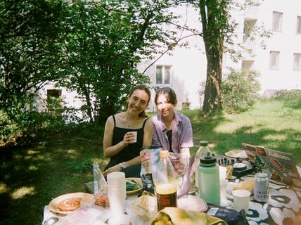 Two people having a picnic in a garden with food, drinks, and a laptop on the table.