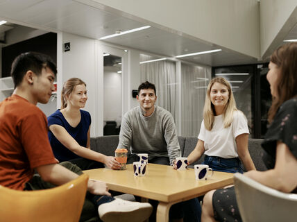A group of five people sit around a table with mugs, having a conversation in a modern room with white walls.