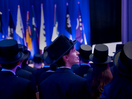 Graduates wearing black hats at a ceremony with flags in the background on a blue-lit stage.
