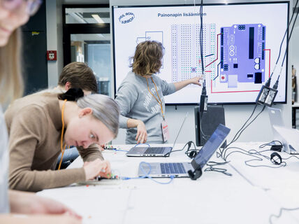 A tech workshop with participants working; a tutor points at a diagram on a screen. Laptops and cables are on the table.