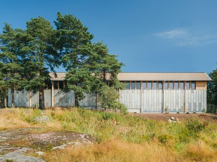 Modern storage building with grey facade with wooden details and windows surrounded by trees and grass under a clear blue sky.