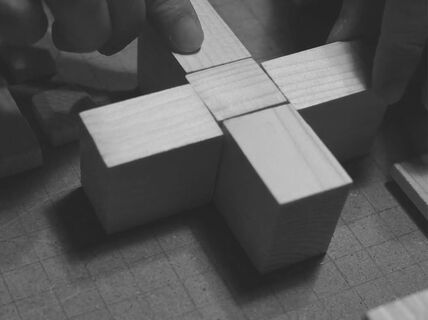 Hands assembling wooden blocks into a cross shape on a patterned surface.