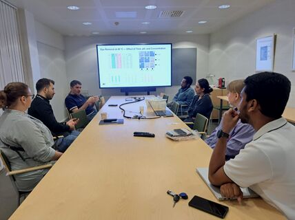 A group of people having a meeting around a long wooden table with a presentation on a large screen.