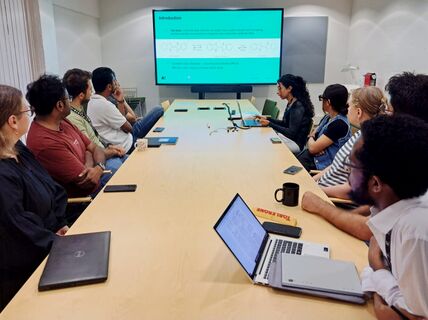 A group of people are seated around a long table, attentively watching a presentation on a large screen.