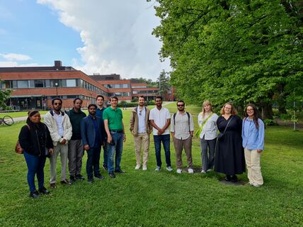 Group of people standing on grass with buildings and trees in the background.
