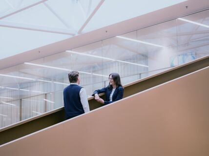 Two people talking on a modern staircase with a glass wall and ceiling lights in the background.