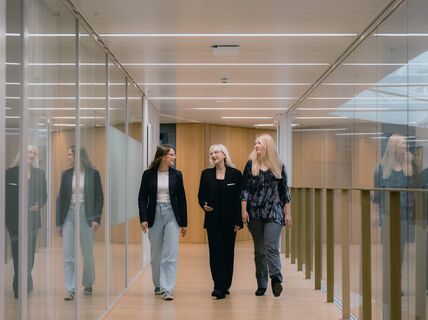 Three women walking through a modern corridor with glass walls, dressed in casual and business attire.