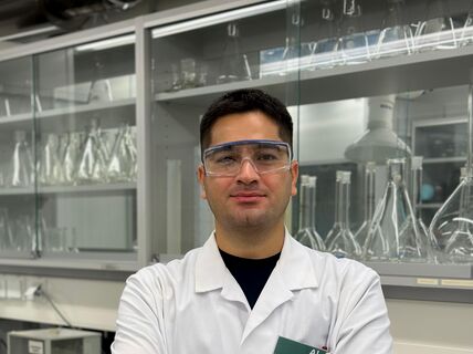 A person in a white lab coat stands in front of laboratory shelves filled with glass containers.