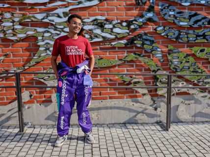 Hitesh Monga wearing Tutor in Aalto University shirt and overalls, standing in front of a brick wall with metal artwork