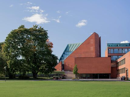 Modern red brick building with glass windows next to trees and a lawn, under a blue sky.