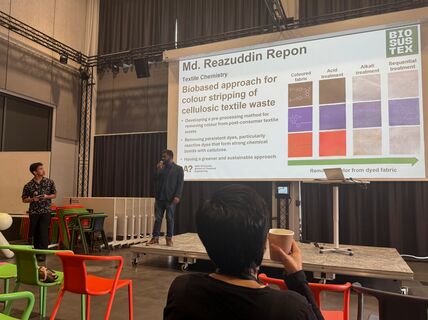 A presentation on biobased colour stripping of textile waste. Two men are speaking on stage. Audience member holding a cup.