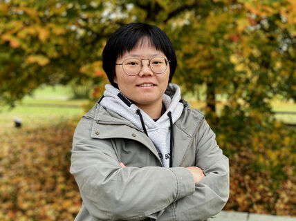 Person standing outdoors in autumn, wearing a grey hoodie and green jacket. Trees in the background with orange leaves.