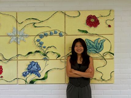 Nainen seisoo kukkamaalauksen edessä, vaalealla taustalla, kädet ristissä.