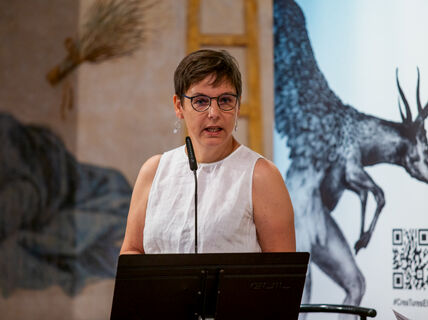 Person in a white blouse speaks into a microphone, presentation slides projected on the wall behind.