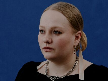 Person with blonde hair in a ponytail, wearing a black dress, silver necklace, and earrings with a blue backdrop.