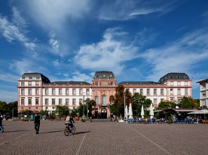 A large, historic building with a pink facade and grey roof in a square with people walking and cycling.