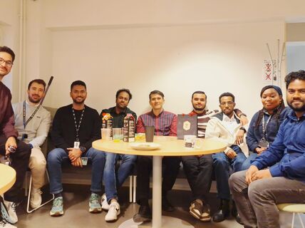 A group of eight people is sitting around a table with drinks and snacks. They are indoors with a bright background.