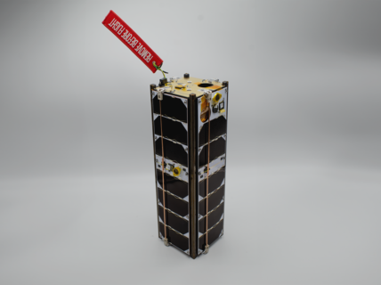 Pieni satelliitti mustilla paneeleilla ja punaisella lapulla, jossa lukee 'REMOVE BEFORE FLIGHT', harmaalla taustalla.