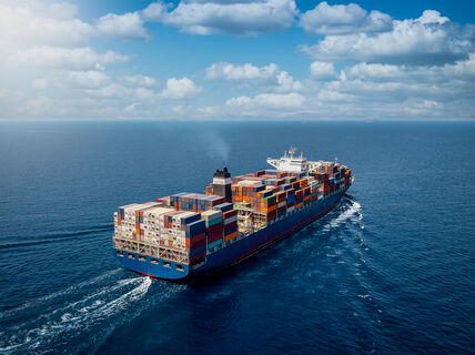 A large cargo ship loaded with colourful containers sails across the blue ocean under a partly cloudy sky.