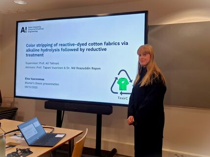 A woman presenting her master's thesis on textile chemistry at Aalto University. A laptop and notes are on the table.