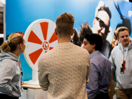 People gathered around a red and white spinning wheel at an event. A blue backdrop with a logo is visible.
