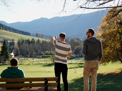 Kolme ihmistä puistossa, taustalla vuoria. Yksi istuu penkillä, kaksi seisoo maisemaa katsellen.