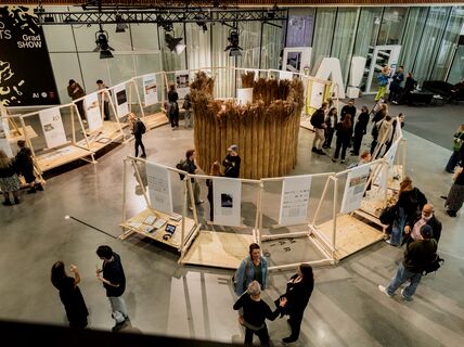 People at an art exhibition with wooden structures displaying artworks. A circular installation made of tall reeds is in the centre.