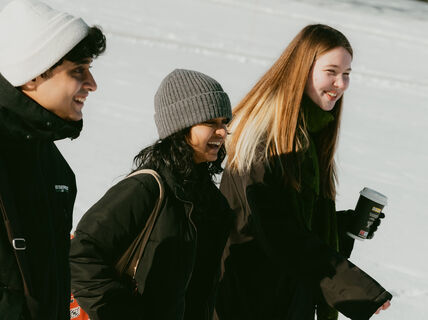 Three people in winter clothing walking outdoors on a snowy day. One holds a coffee cup.