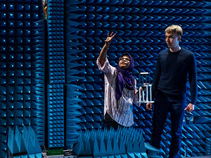 Kaksi ihmistä kaiuttomassa huoneessa, jossa on siniset akustiset vaahtopaneelit. Toinen elehtii, toinen pitää laitetta.