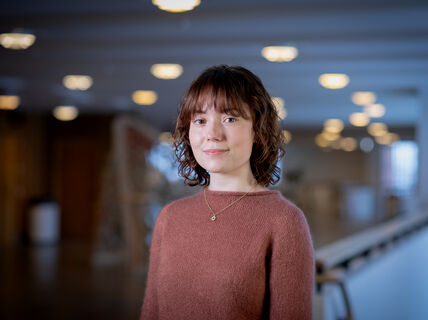 En person med rosa tröja och halsband står inomhus med suddiga ljus i bakgrunden.