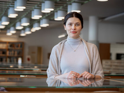 Henkilö seisoo kirjastossa, yllään vaaleanharmaa poolopaita ja huivi. Taustalla näkyy kirjahyllyjä ja kattovaloja.